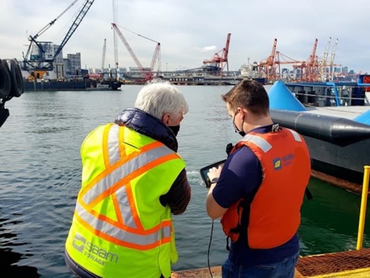 Two men in PPE gear stand dockside in a port looking at the screen of a Deep Trekker DTG3 submersible ROV