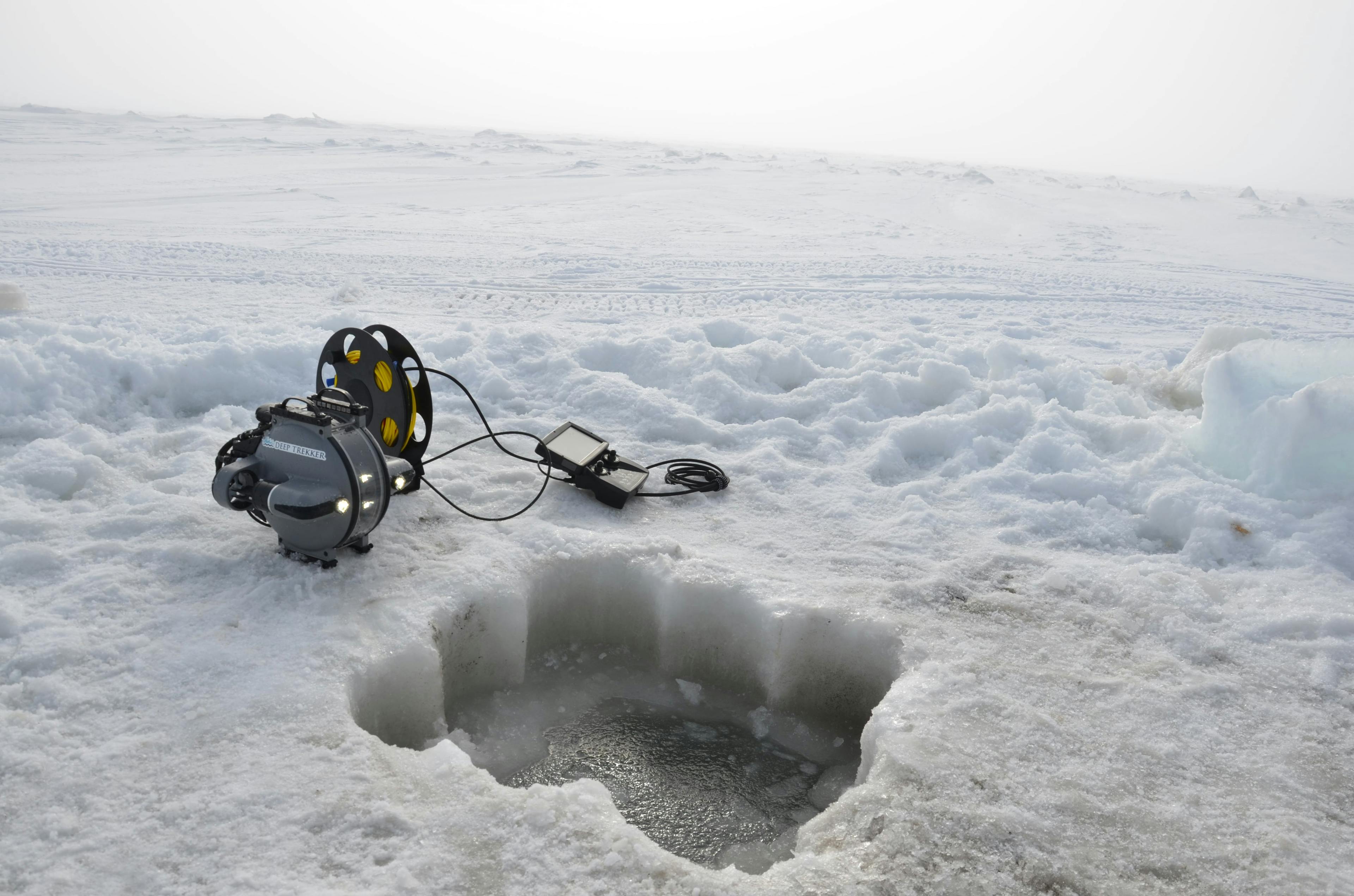 Lagoon covered in ice and snow with a large square drilled into the ice. Next to the hole is a Deep Trekker DTG3 remotely operated vehicle.