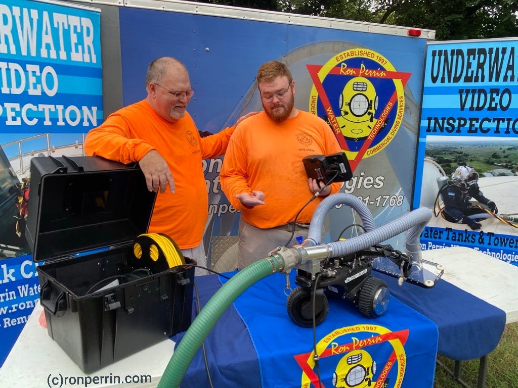 Ron and Robert Perrin stand behind display table with the Deep Trekker DT640 VAC utility vehicle on the table. The table is covered in blue fabric and the Ron Perrin Water Technologies Inc logo. 