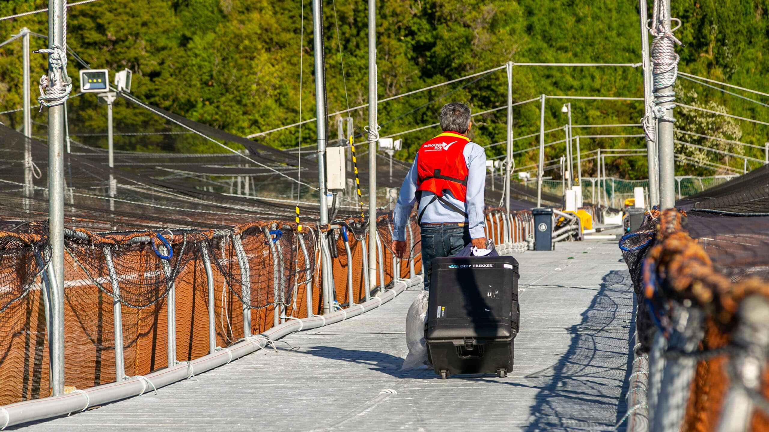 Людина йде по причалу рибної ферми, везучи пластиковий кейс на колесах з Deep Trekker DTG3 ROV.