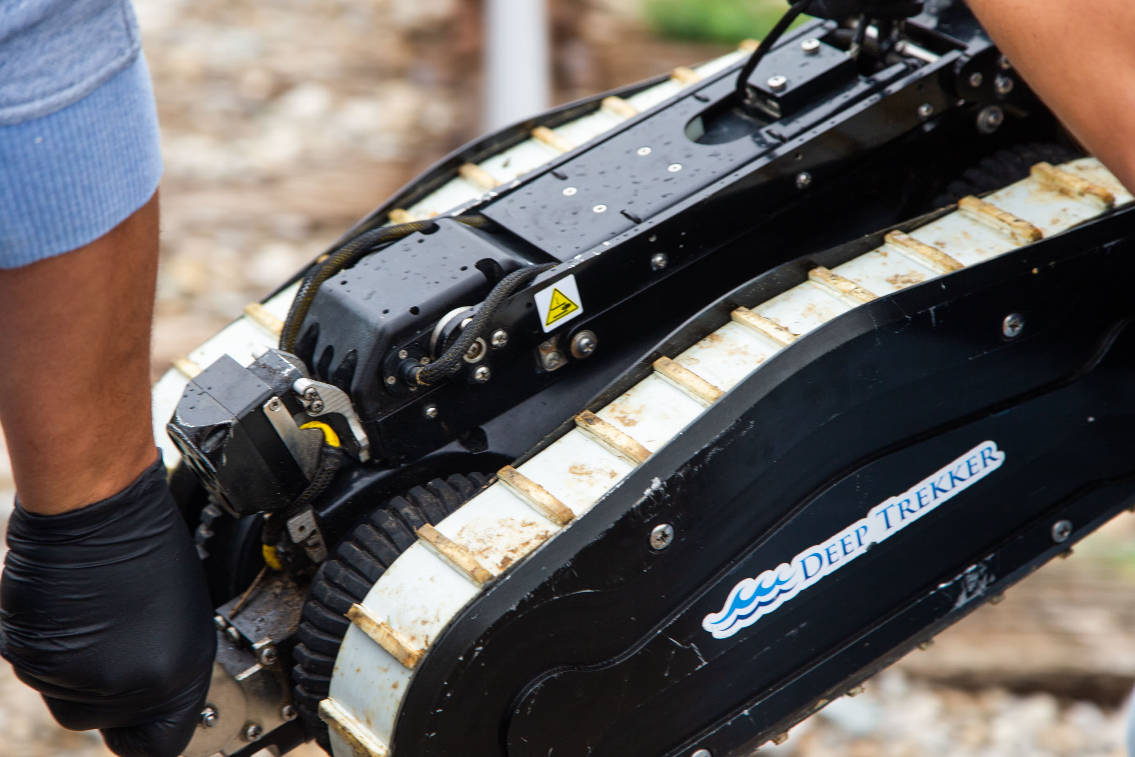 Deep Trekker's DT340 pipe crawler with a blue and white wave logo is being held above muddy ground by a man wearing black gloves and a blue sweater