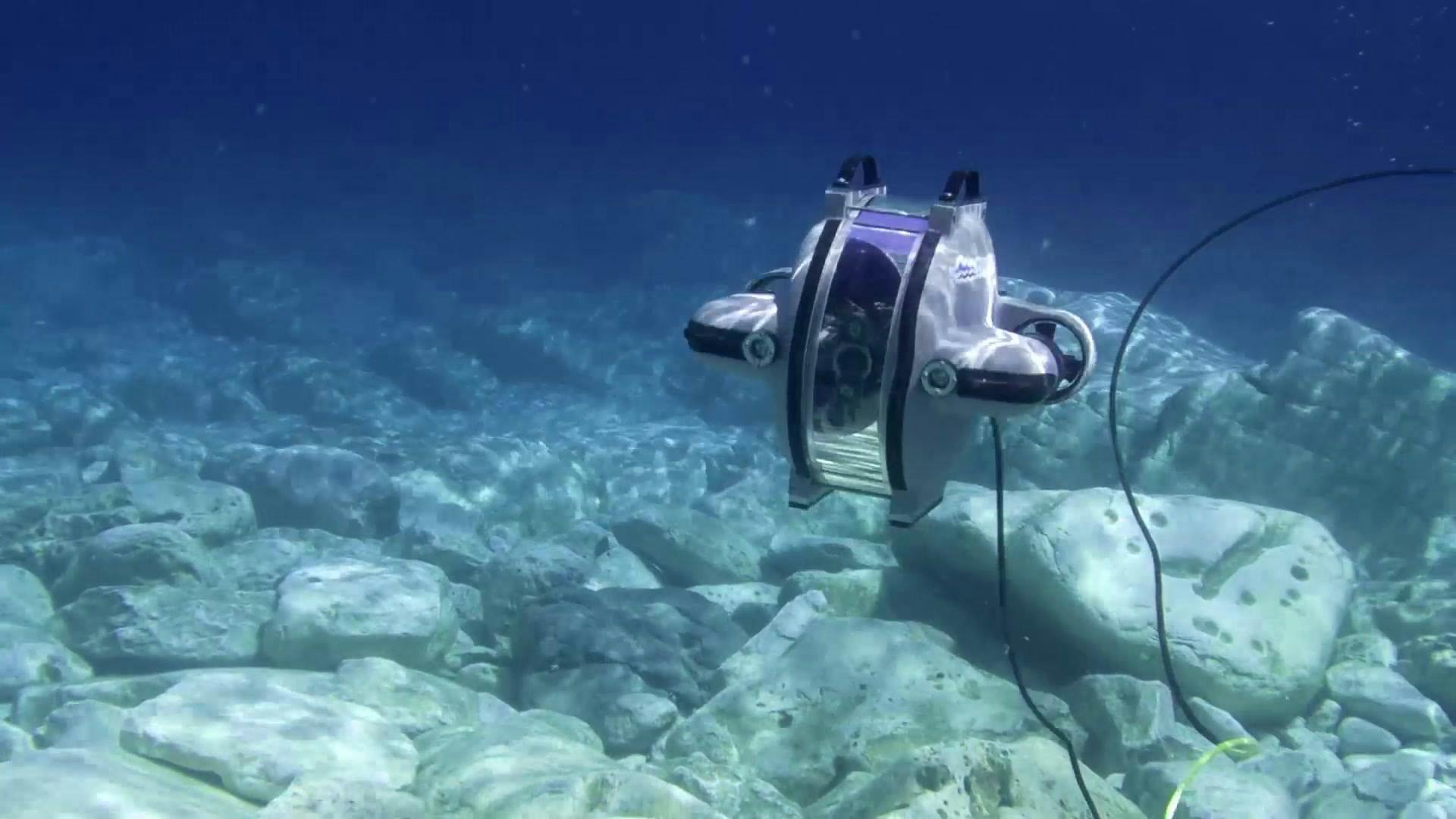 Underwater image of Deep Trekker DTG3 ROV swimming over big rocks in clear, blue water