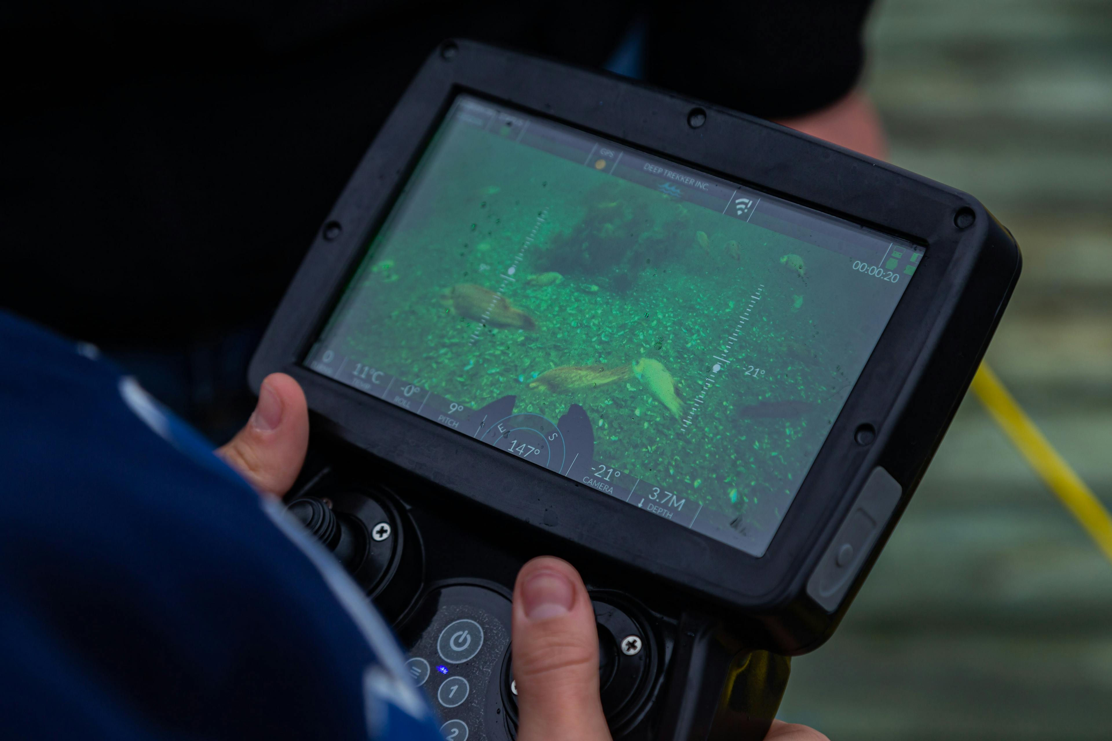 Close up view of the handheld controller screen of a Deep Trekker DTG3 vehicle showing an underwater view of fish and the bottom of a lake. 